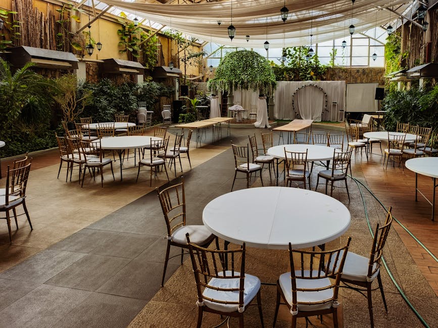 An empty wedding venue setup with round tables and chairs indoors, surrounded by plants.